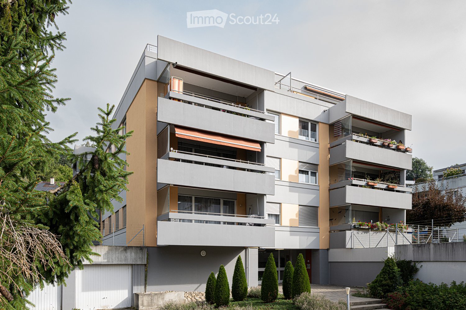 Modern multi-story apartment building with balconies, grey and beige facade, surrounded by greenery and trimmed trees.