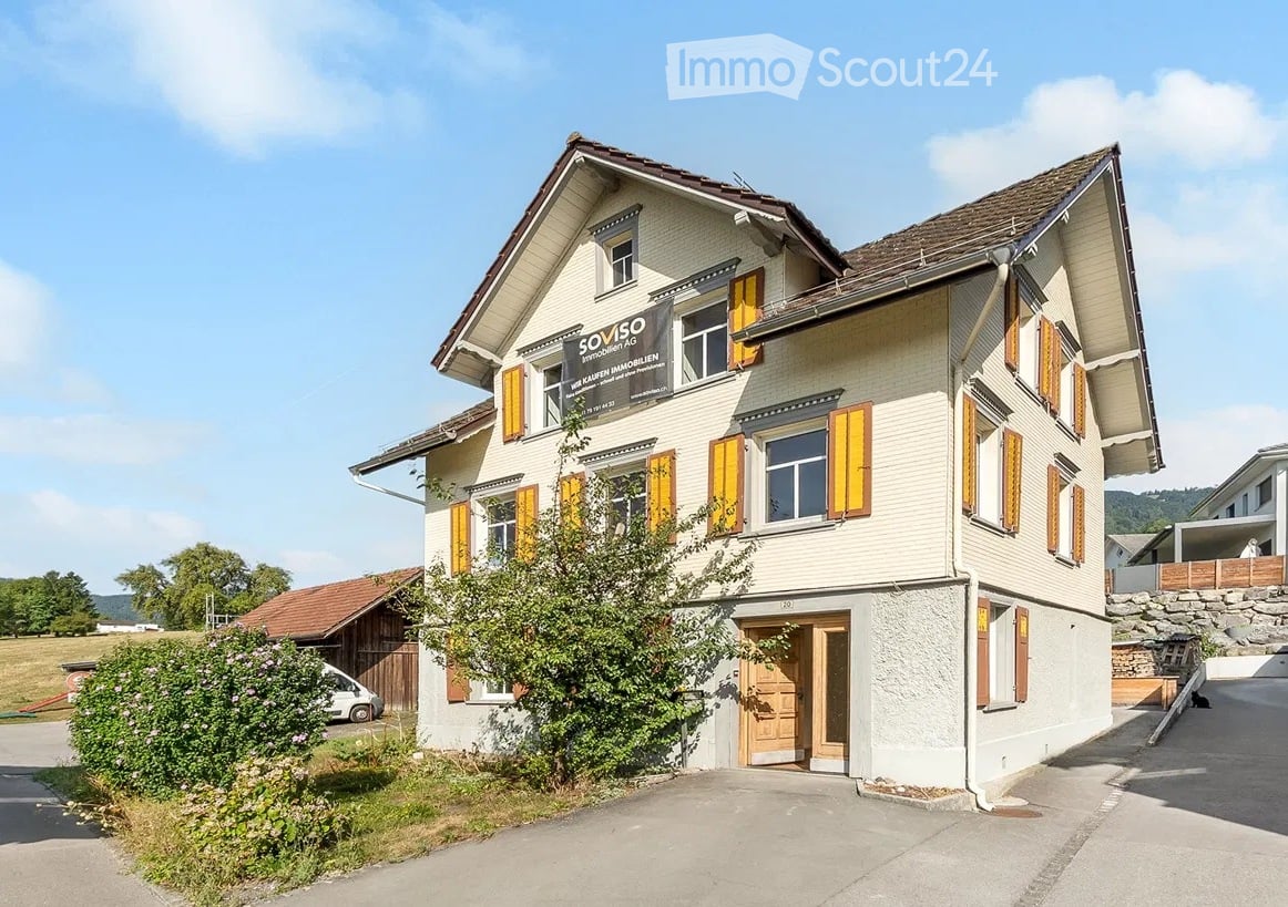 Two-story house with light-colored walls, yellow shutters, and a brown roof. The house has multiple windows and a central entrance with a wooden door. There is a small garden area in front of the house, and a sign on the wall indicates it is for sale.