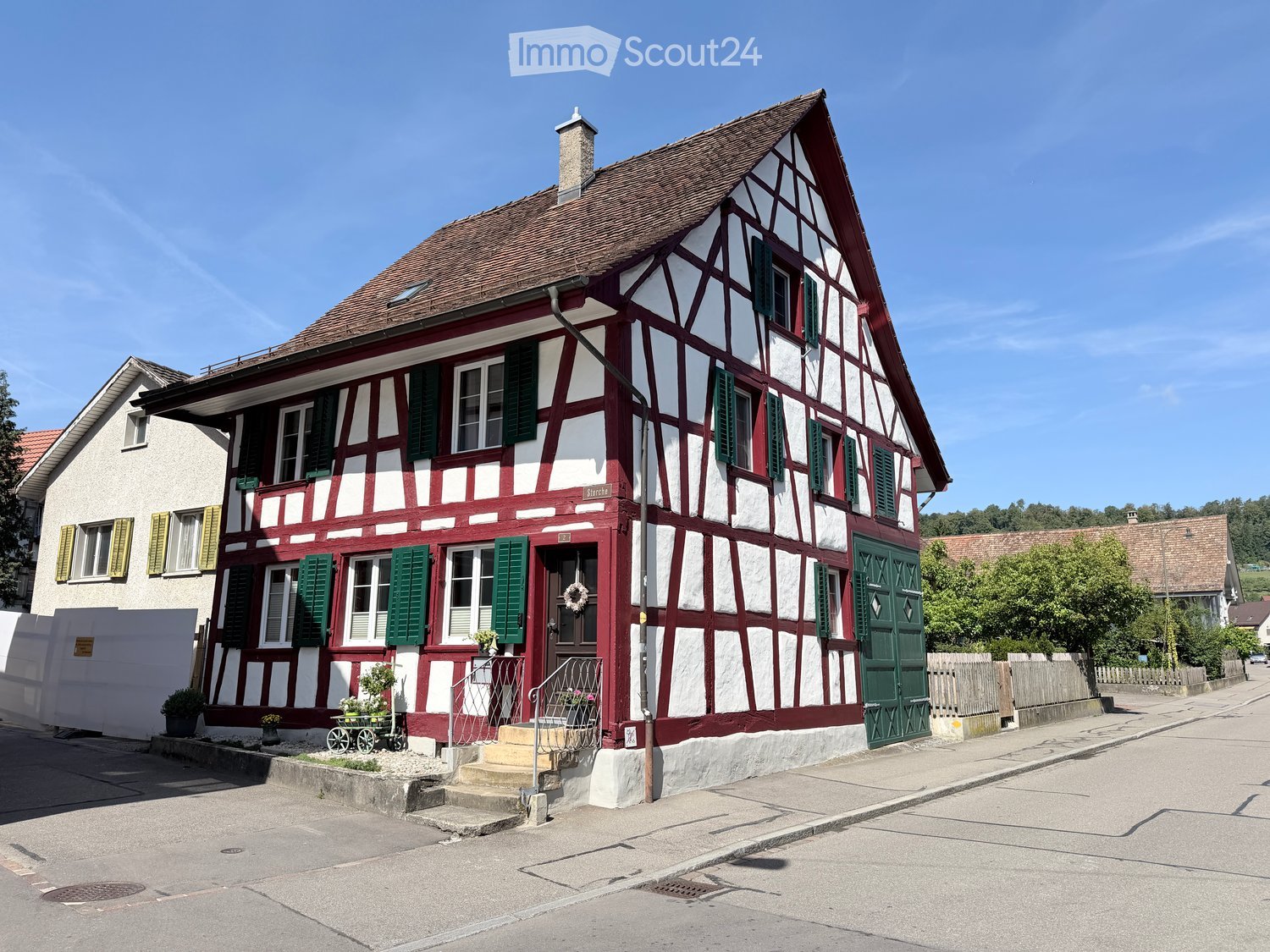 Traditional half-timbered house with red and white exterior, green shutters, and a small garden area with potted plants near the entrance.