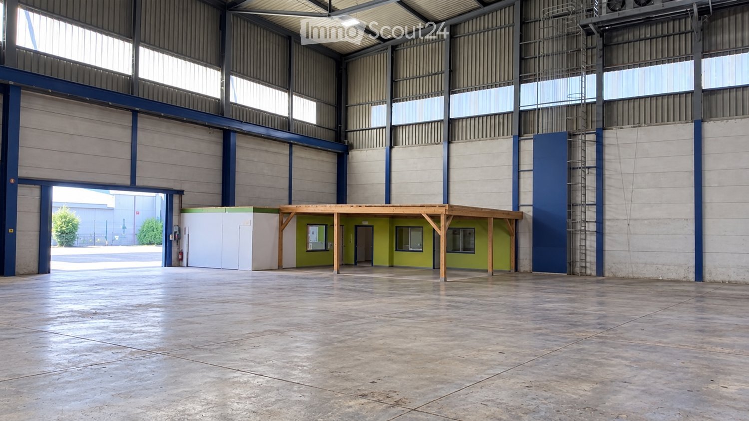 Large industrial warehouse with high ceilings, metal walls, and large windows. Features a wooden platform structure and a small green office area.