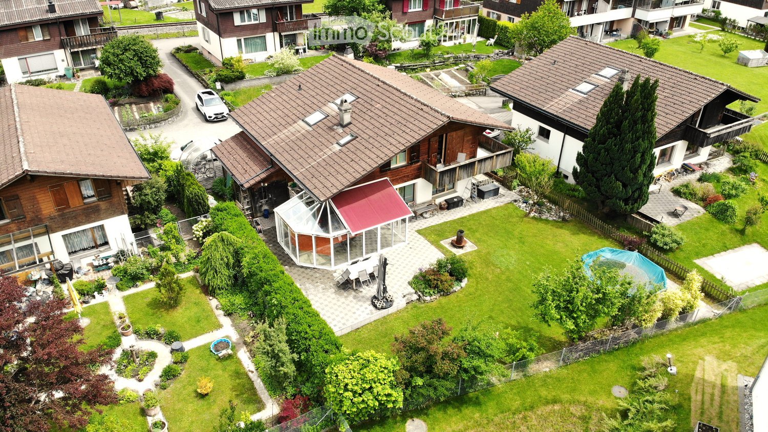 Aerial view of a residential area with multiple houses, well-maintained gardens, a swimming pool, and outdoor seating areas.