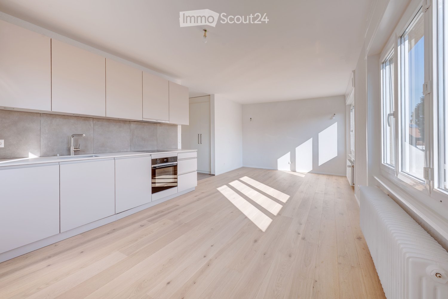 Modern kitchen with white cabinets, light wooden flooring, and large windows allowing natural light to fill the space. Features include a sink, faucet, and built-in oven.