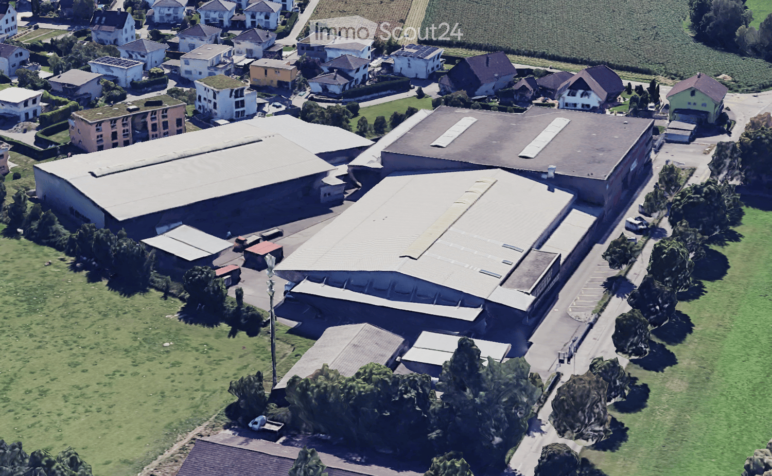 Aerial view of a residential and industrial area with multiple buildings, including houses, warehouses, and a large industrial complex with a prominent roof structure. The area is surrounded by green fields and trees.