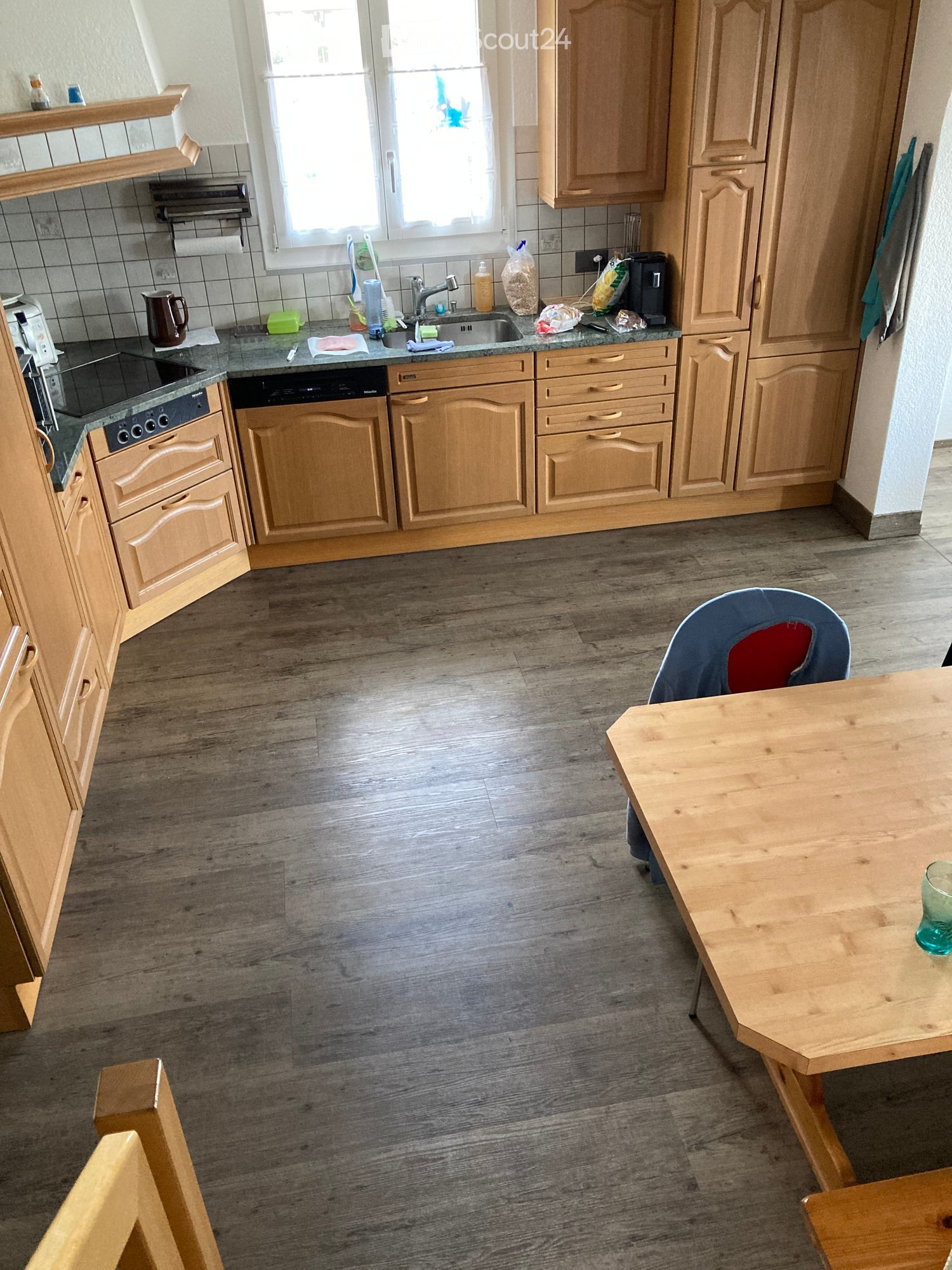 The kitchen features light wooden cabinets, a granite countertop, and a wooden dining table with a single chair. The floor is made of wood, and the sink area includes a faucet and various small items.