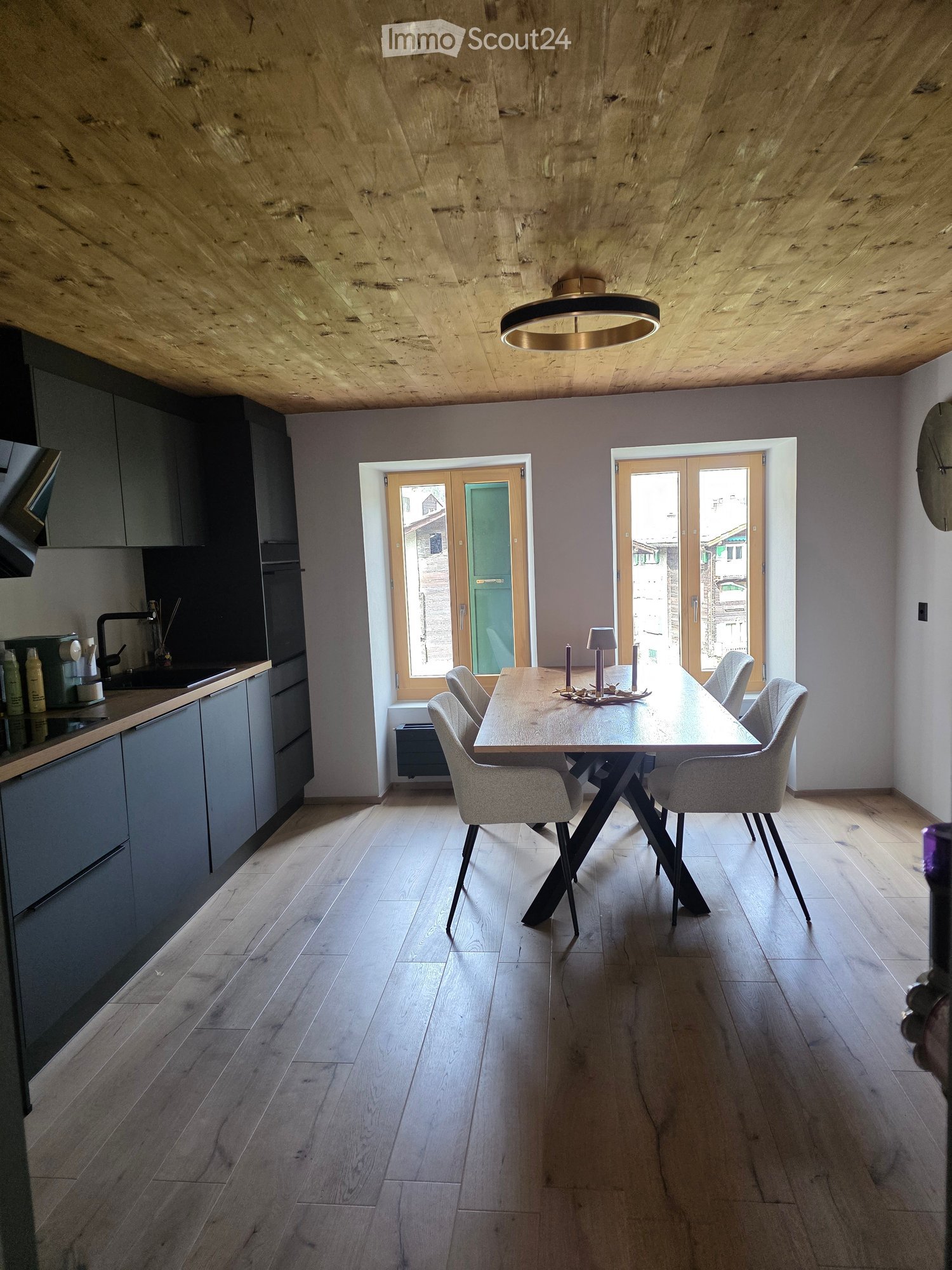 Modern dining area with a wooden dining table and chairs, grey kitchen cabinets, and a sink with a faucet. The room features wooden flooring and a wooden ceiling with a pendant light.