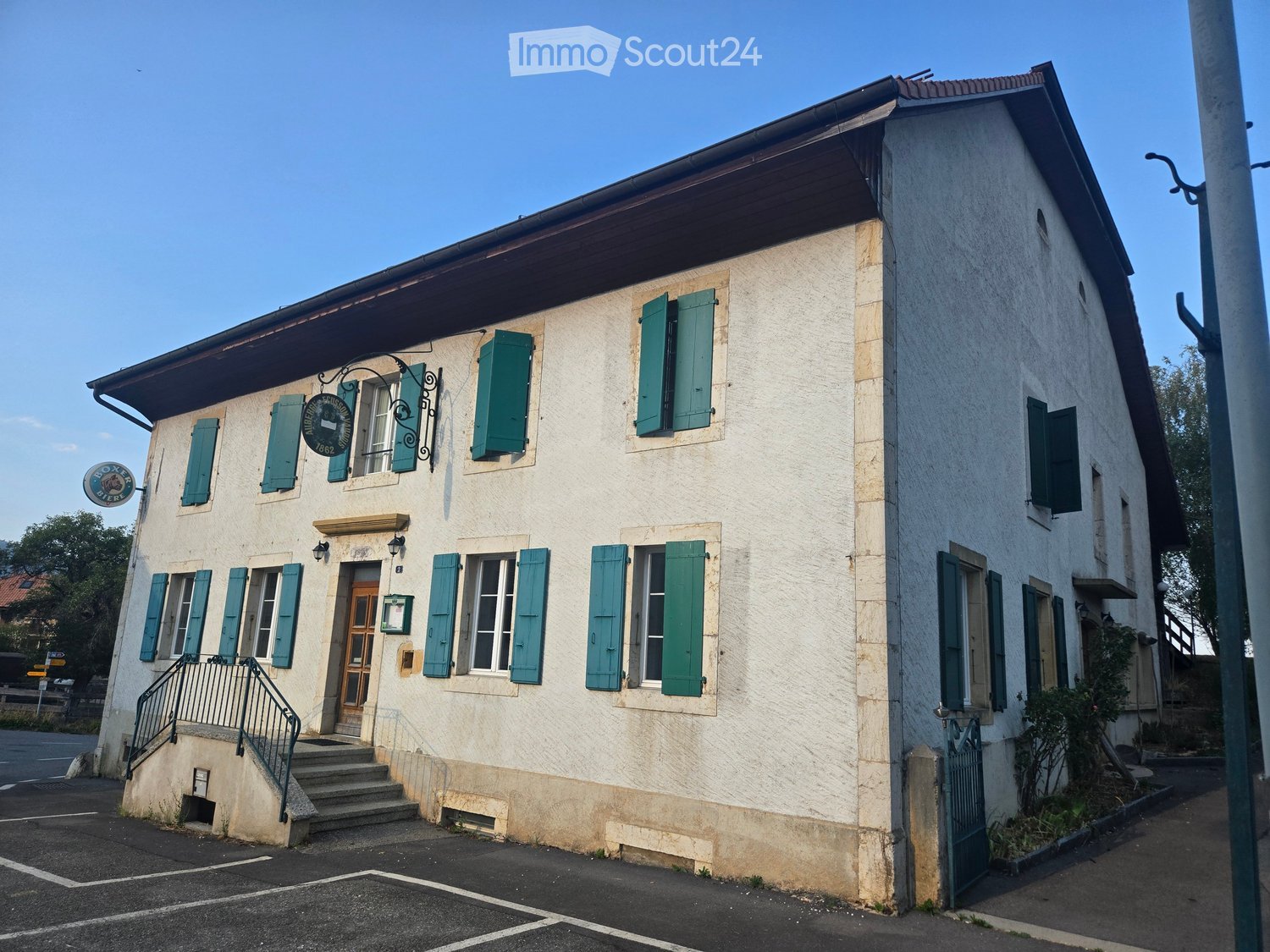 Two-story building with light-colored walls, green shutters, and a small staircase leading to the entrance. The building has a sign indicating "Boxe" and the year 1862.