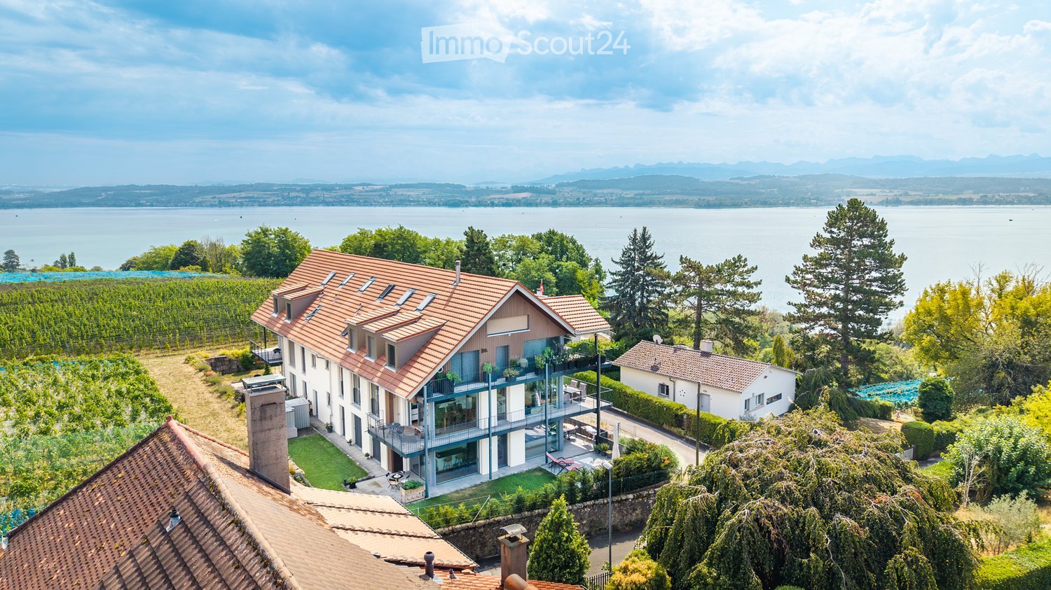 A modern two-story house with a red-tiled roof, large windows, and a spacious balcony overlooking a scenic lake and vineyard area.
