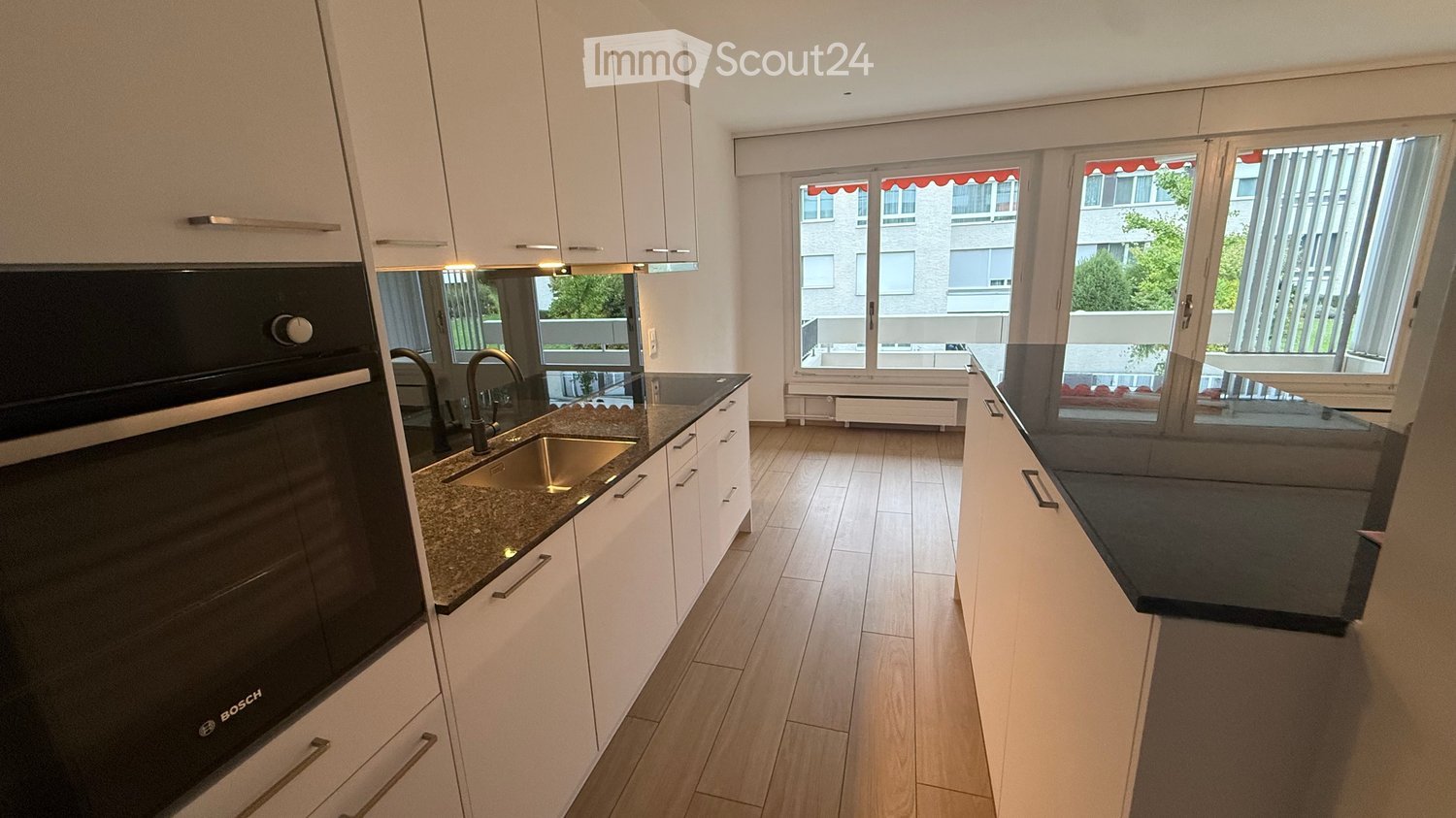 Modern kitchen with white cabinets, granite countertops, stainless steel sink, and Bosch oven. Large windows provide natural light and a view of the balcony.