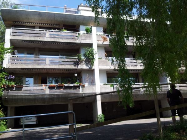 Modern multi-story apartment building with balconies, greenery, and a covered parking area.