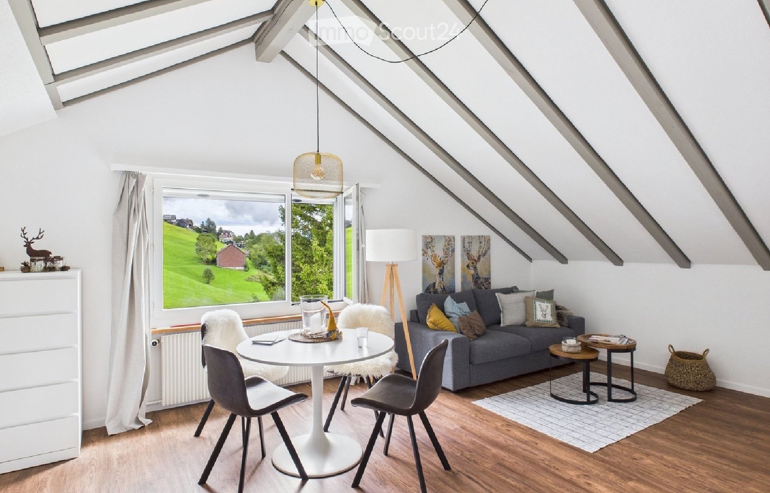 Bright attic living room with exposed wooden beams, large window offering a scenic view, grey sofa, white dining table, modern chairs, and decorative items.