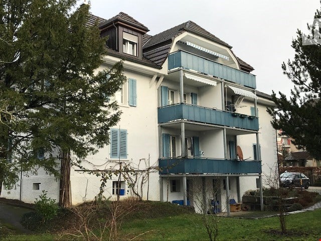 A multi-story residential building with light blue shutters, balconies, and a well-maintained lawn.