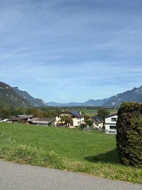 A peaceful rural landscape with a cluster of houses, green fields, and distant mountains under a clear blue sky.