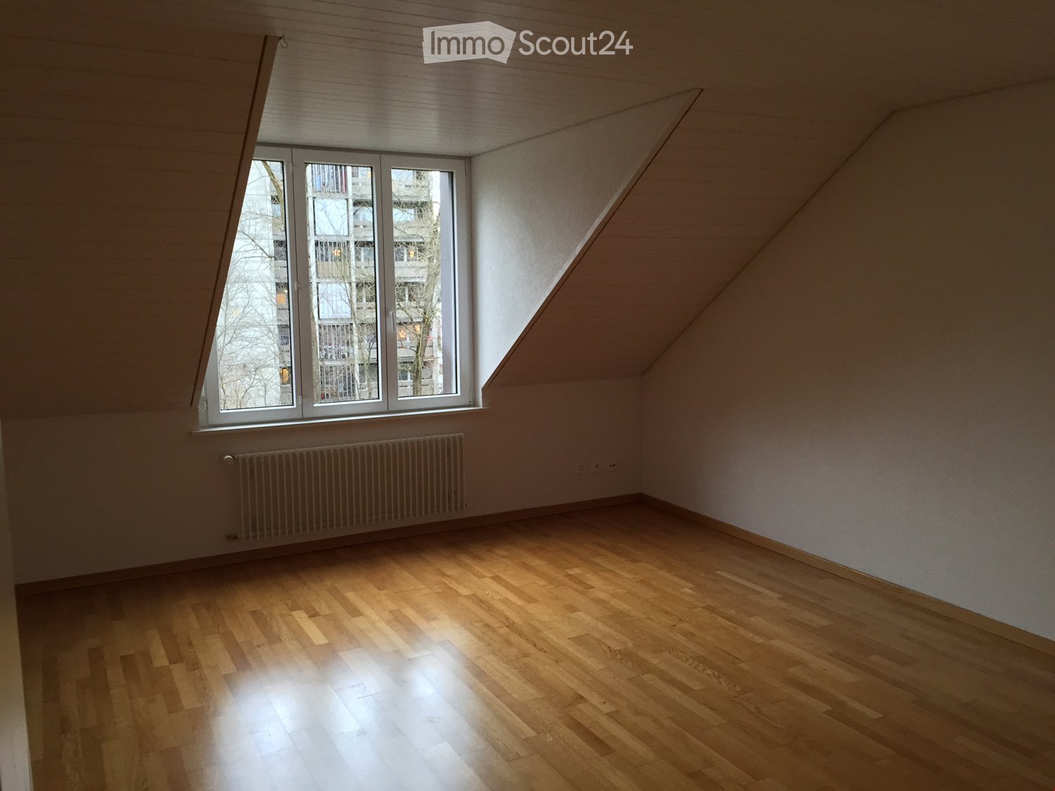 Empty attic room with wooden flooring, white walls, and a large window letting in natural light. The room features a slanted ceiling and a radiator below the window.