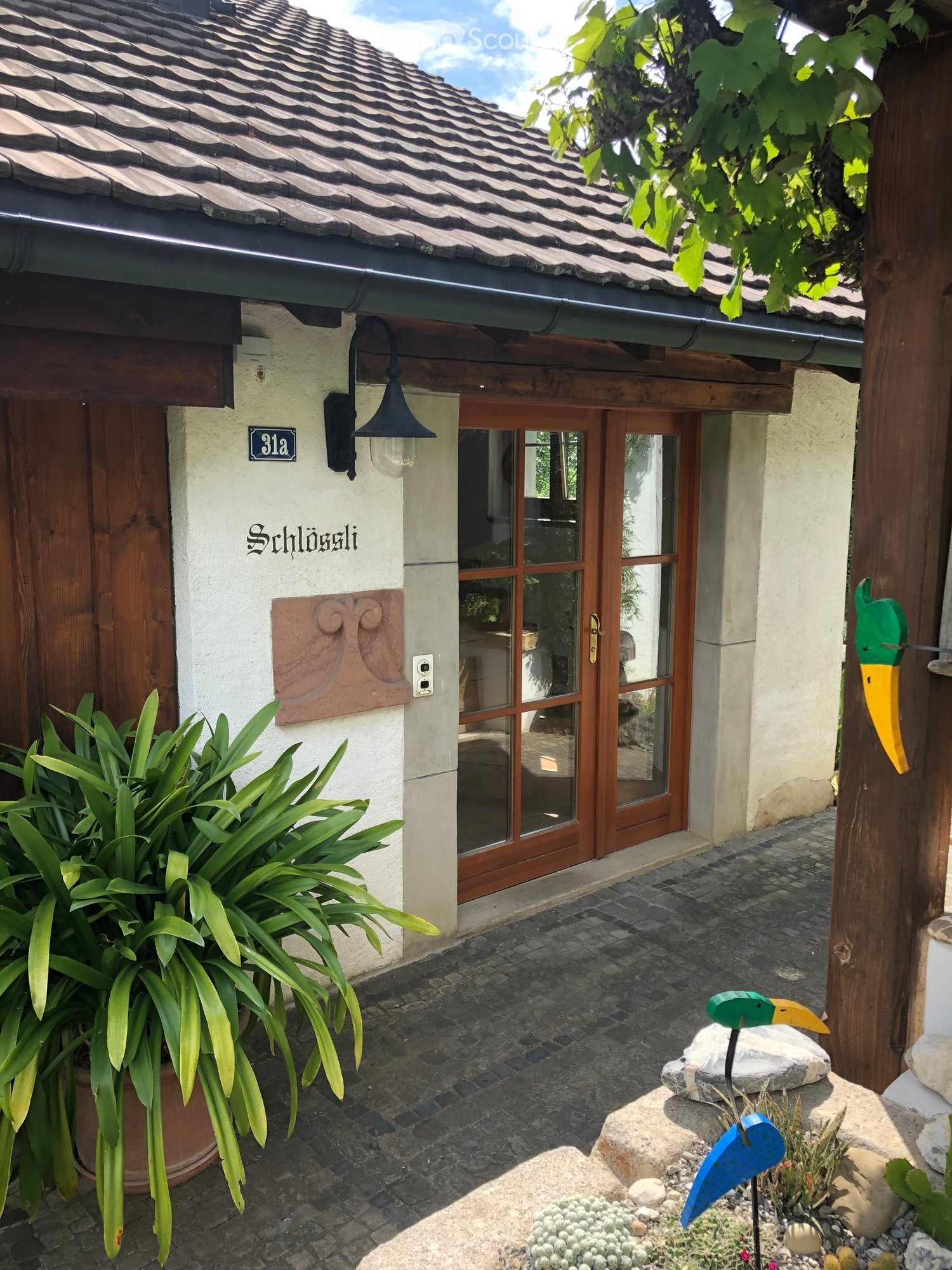 Entrance to a house with a decorative sign, a wooden door, glass-paneled double doors, and a small garden area with potted plants and decorative bird sculptures.