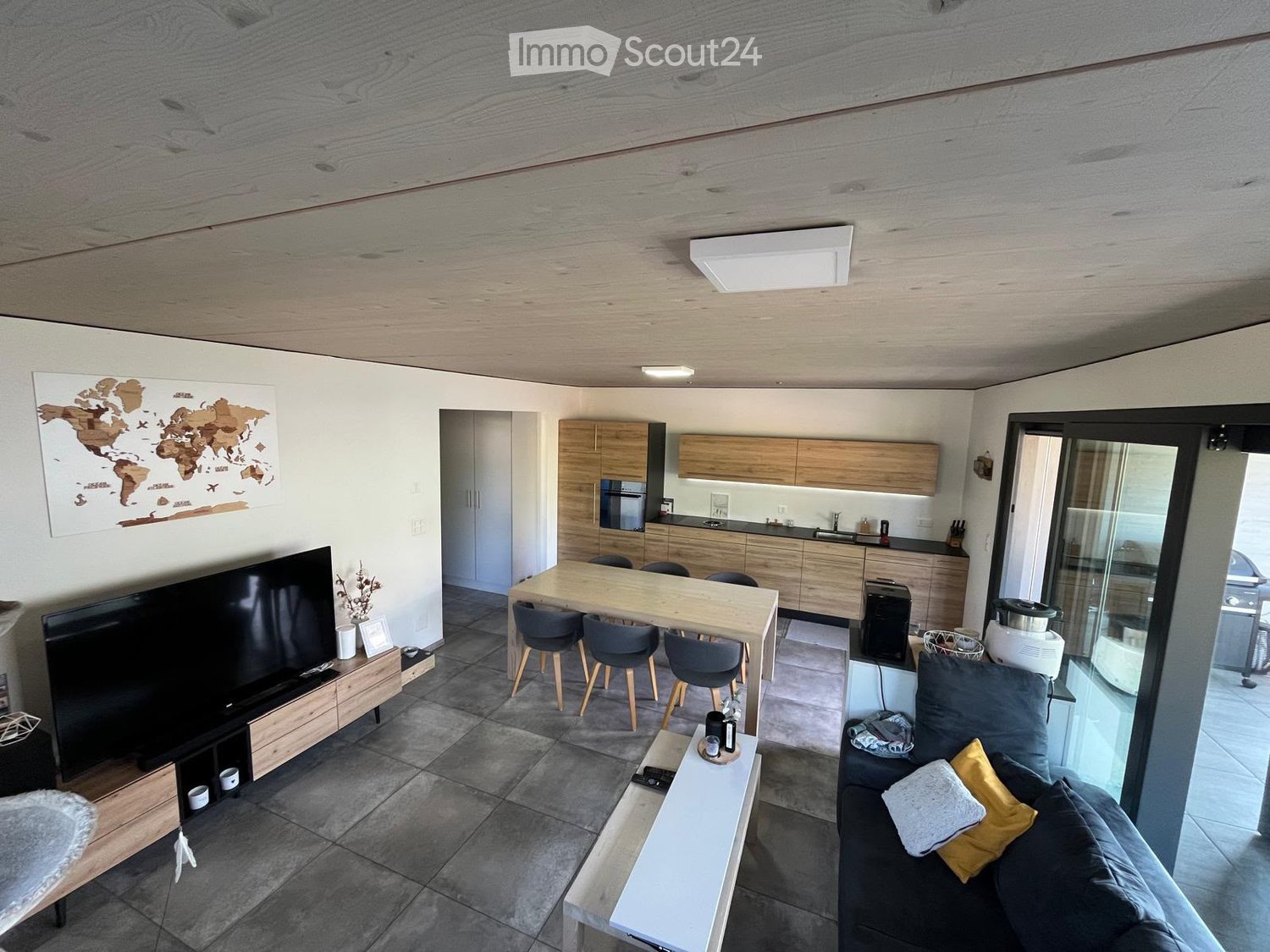 Open-plan living and dining area with a grey sofa, coffee table, TV, dining table with chairs, and a modern kitchen with cabinets and appliances.