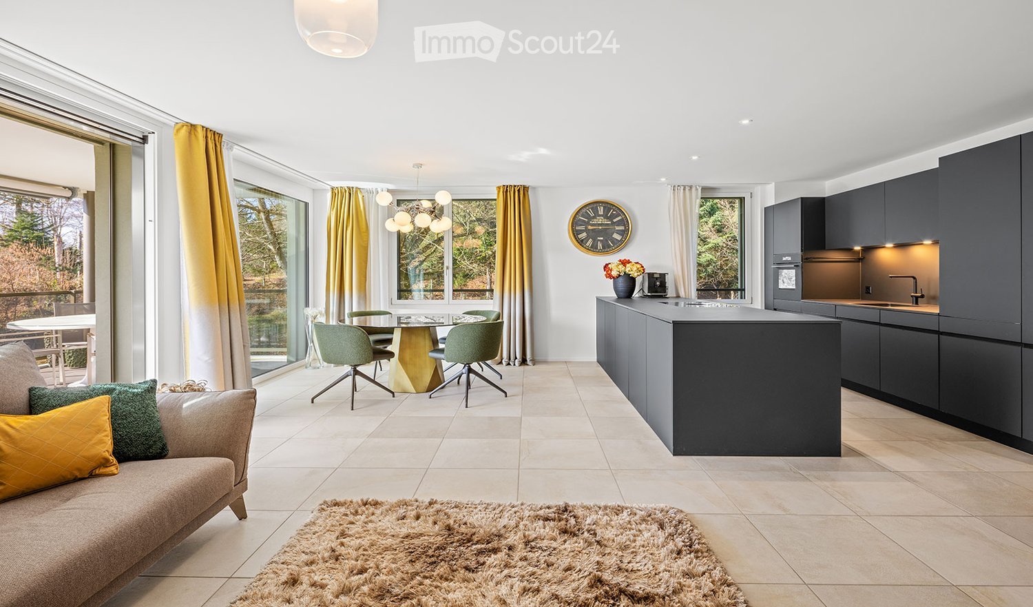 Open-plan living and dining area with a modern kitchen. Features a sofa with cushions, a coffee table, a dining table with chairs, a kitchen island, and fitted kitchen cabinets.