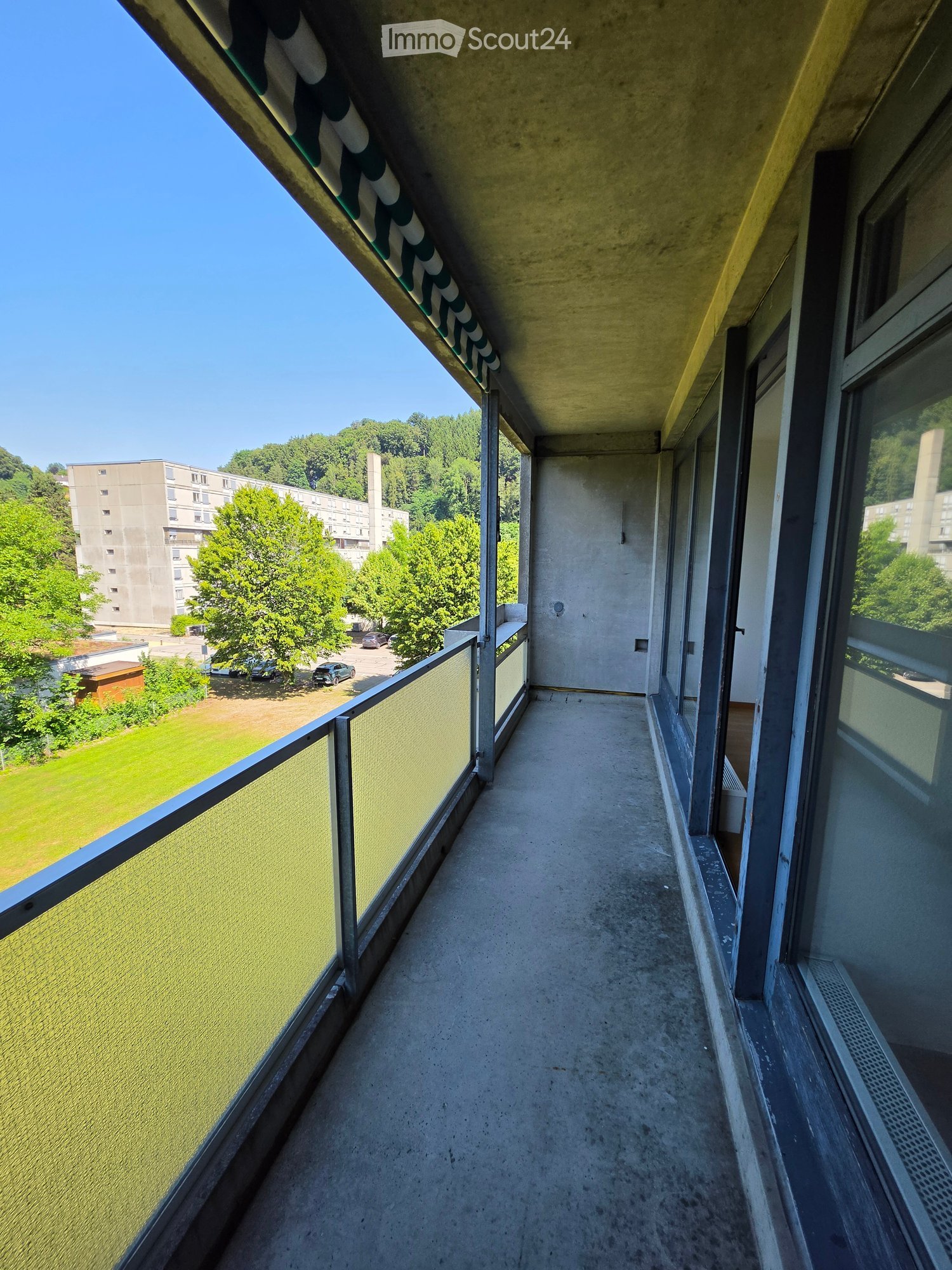 Balcony with concrete floor, glass railing, and view of green park and buildings.