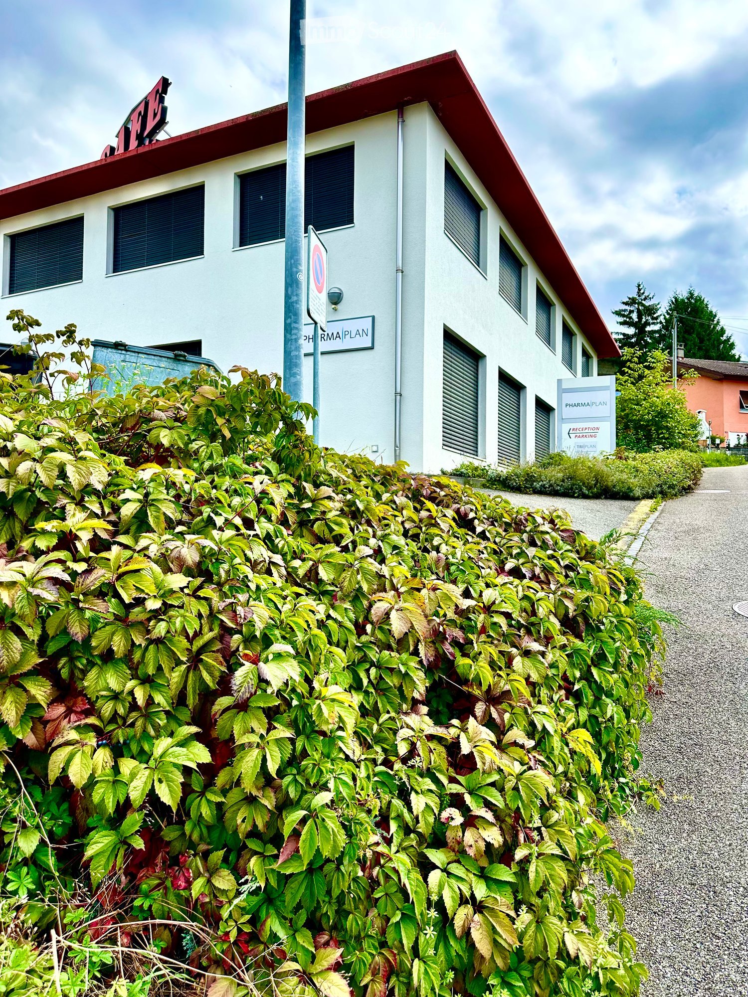 Two-story building with a red roof and white walls, surrounded by greenery and a gravel pathway.