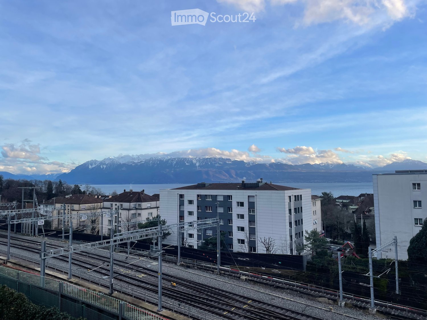 Modern multi-story residential building with white and grey facade, located near train tracks with a scenic mountain and lake view in the background.