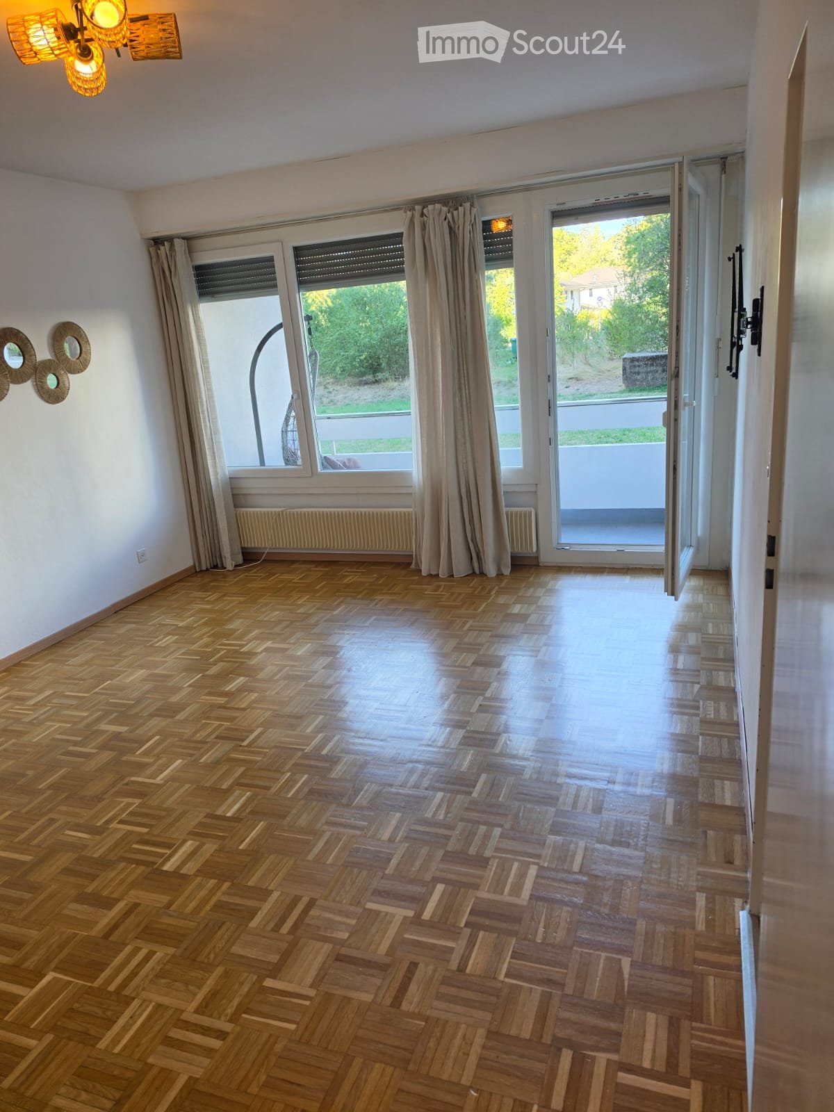 Empty room with wooden herringbone flooring, large windows with curtains, and a sliding glass door leading to a balcony.
