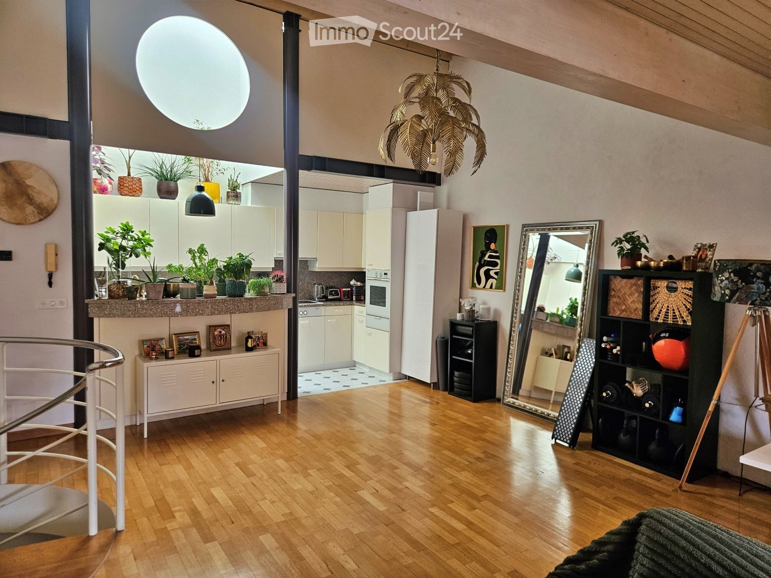 Open-plan living and kitchen area with wooden flooring, white cabinets, a large mirror, and various decorative items including potted plants and artwork.
