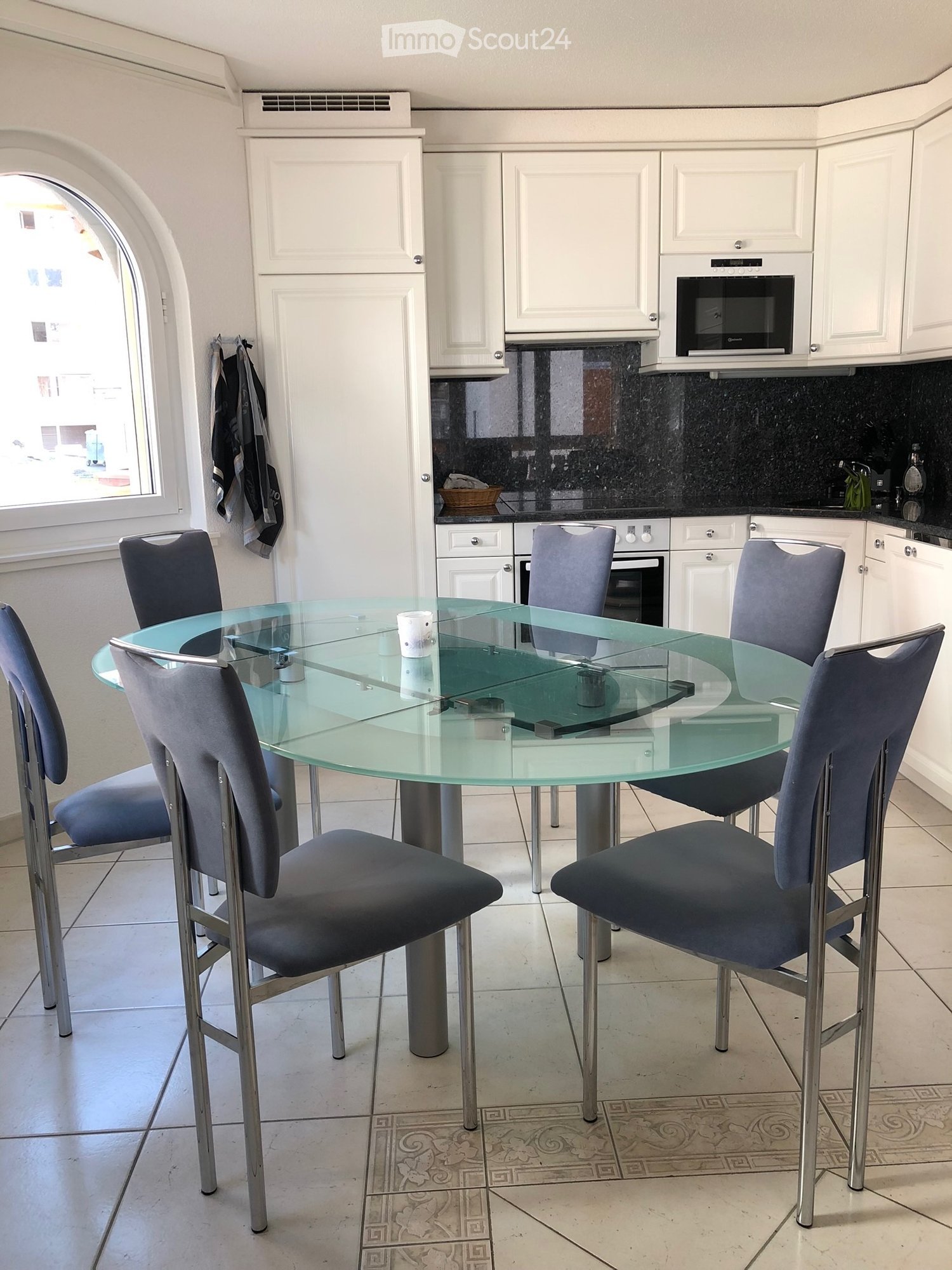 Modern kitchen with white cabinets, a glass dining table surrounded by four grey chairs, and a microwave on the counter.