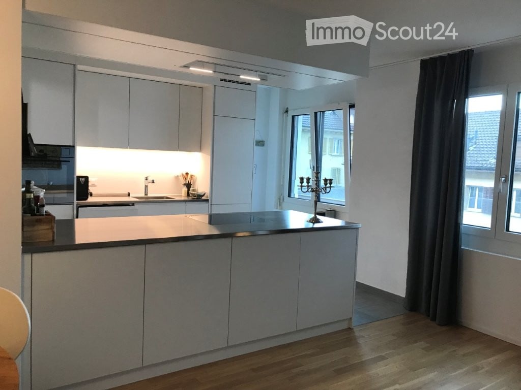 Modern kitchen with white cabinetry, a central island, and a sink area. Features include a candle holder on the island, a window with dark curtains, and built-in lighting.