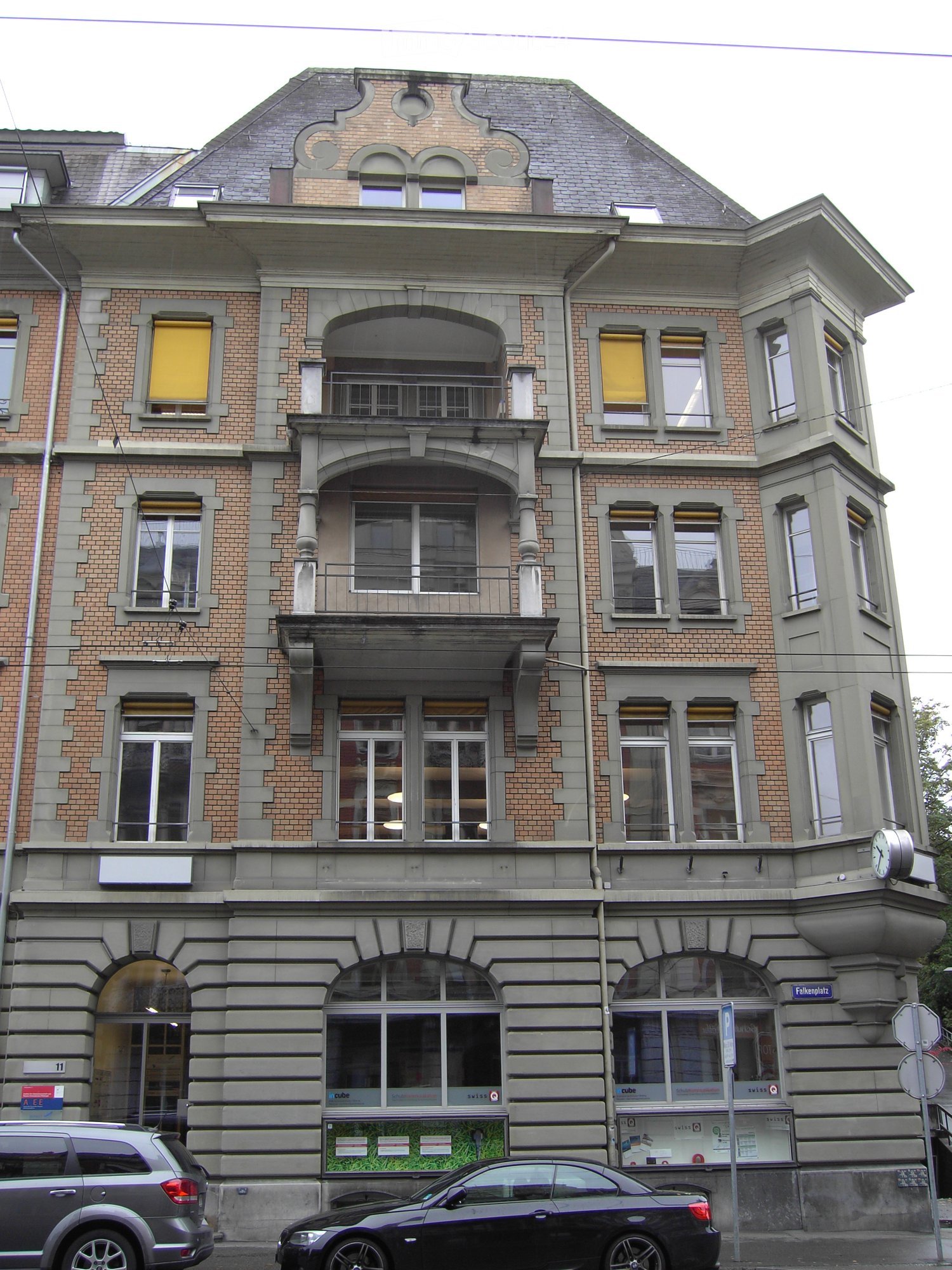 Four-story brick building with arched windows, balconies, and decorative stonework. Features a central entrance with large glass doors and a small green area in front.