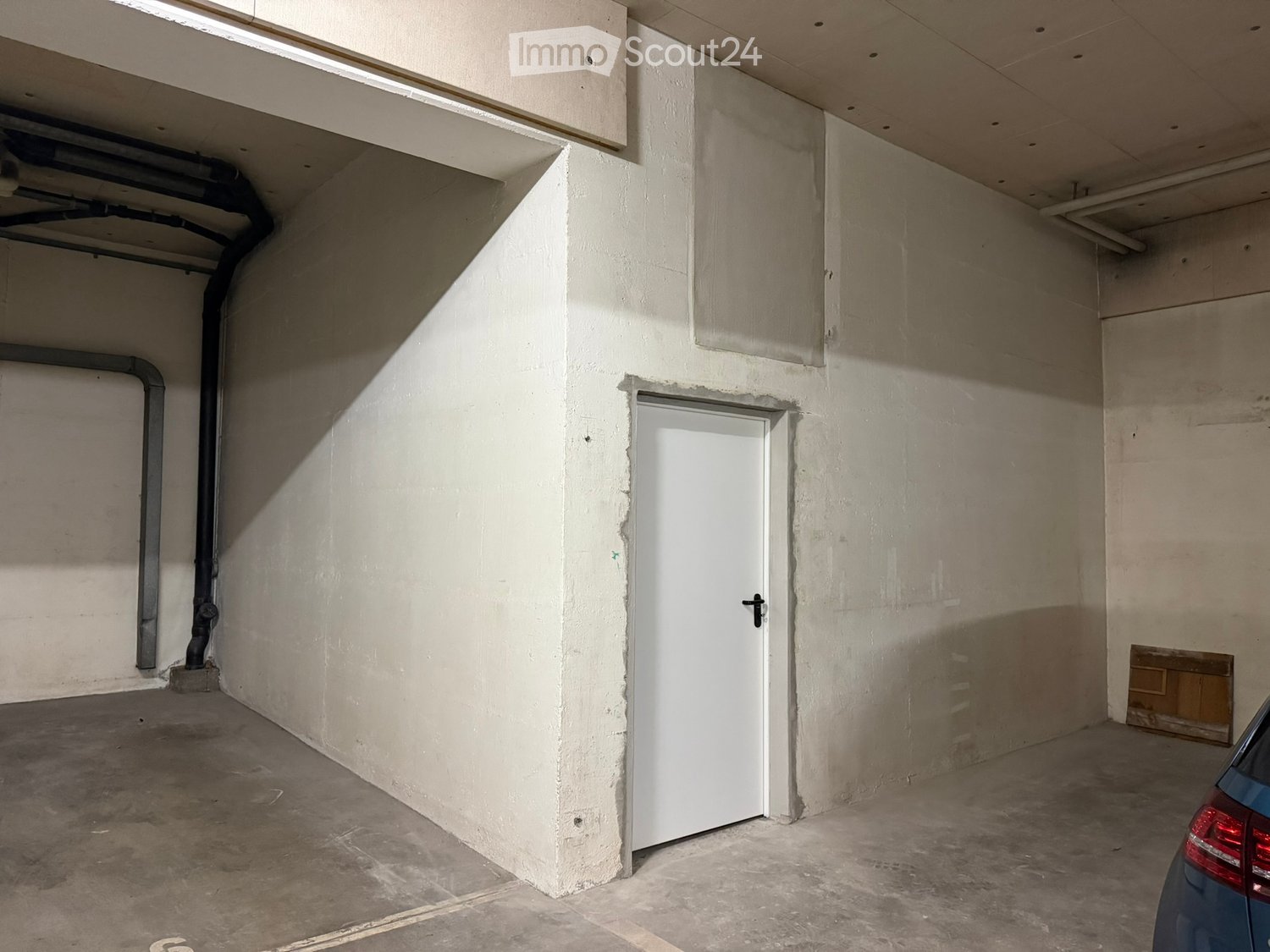 Empty underground parking space with concrete floors and walls, a single white door, and a blue car partially visible on the right side.