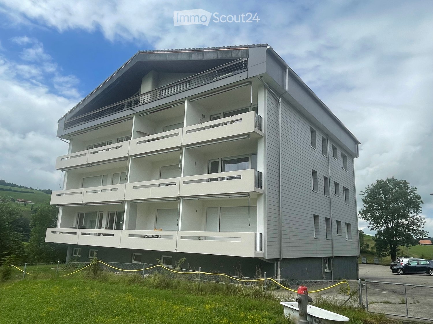 Modern three-story building with multiple balconies, grey siding, and large windows, situated on a grassy slope with a parking area nearby.