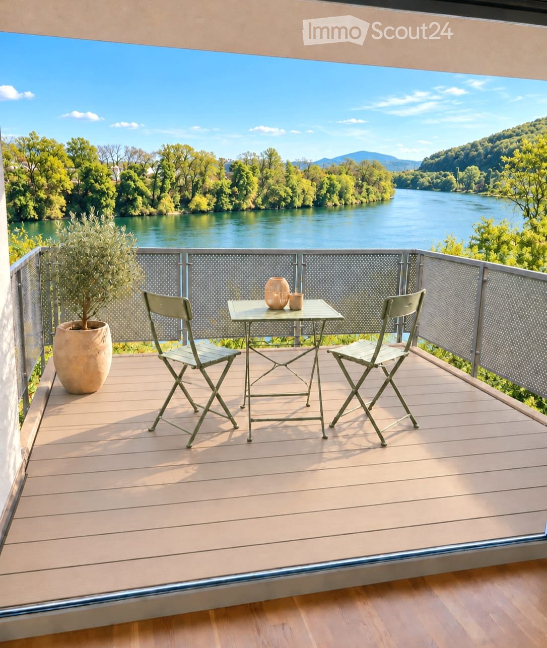 Balcony with wooden decking, metal railing, table with two chairs, vase, and cup, overlooking a river and green landscape.