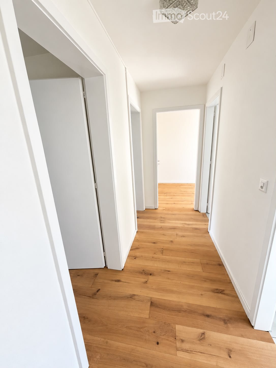 Bright, minimalist hallway with light wooden flooring, white walls, and several doors leading to other rooms.