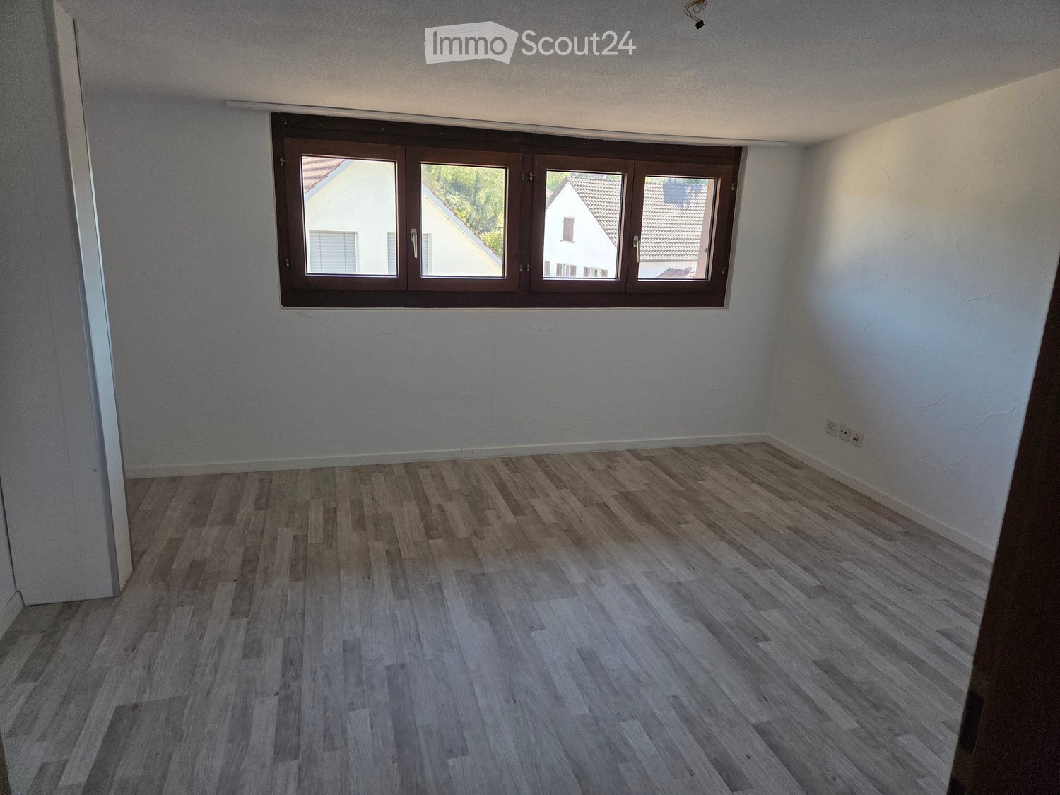 Empty room with light grey walls and wooden flooring, featuring a large window with brown frames and a visible electrical outlet on the right wall.