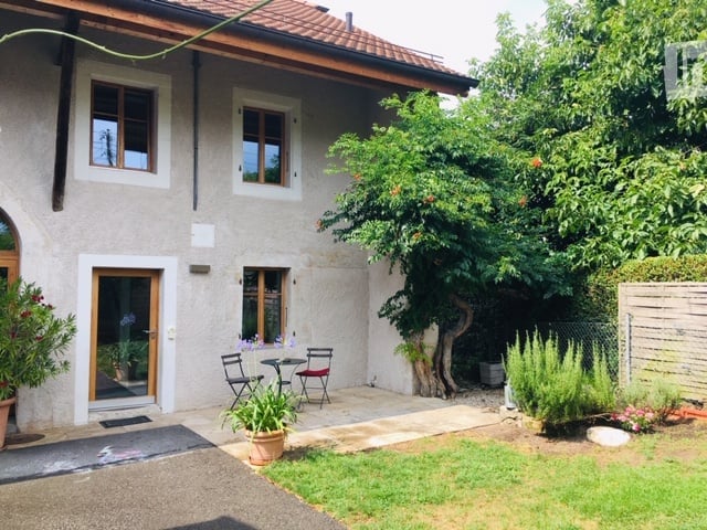 A small, well-maintained garden area adjacent to a house. Features include a patio with a small table and chairs, potted plants, a tree, and neatly trimmed grass.