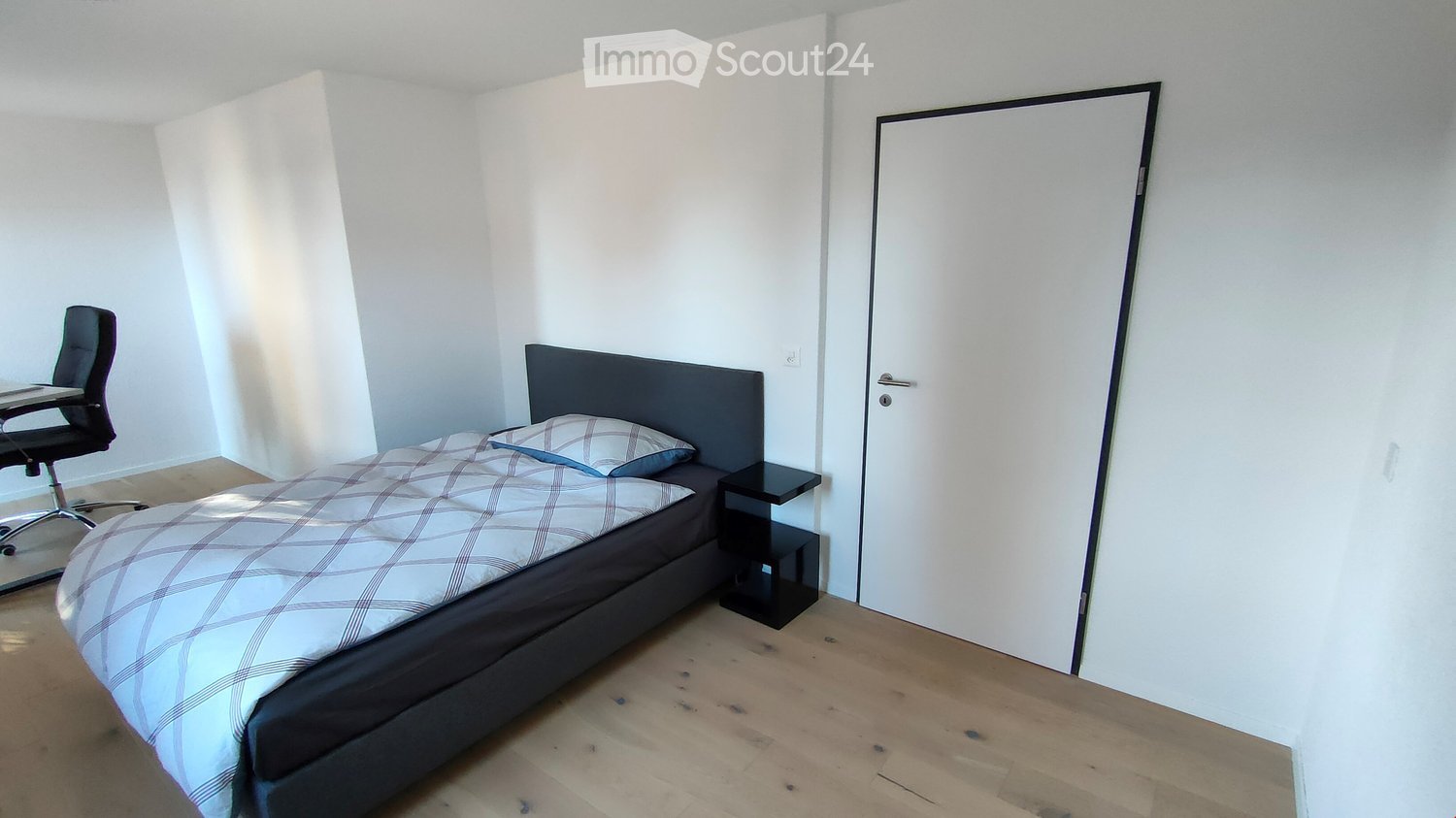 Modern bedroom with a double bed, nightstand, desk, and chair. Light wooden flooring and white walls.