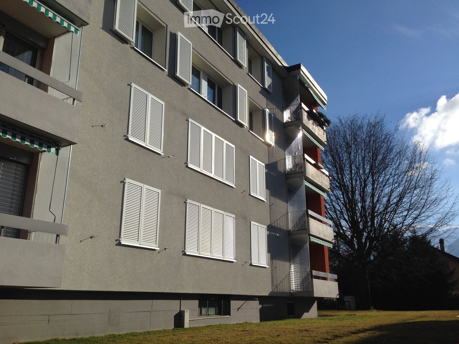 Four-story residential building with grey facade, white shutters, and multiple balconies with green awnings.