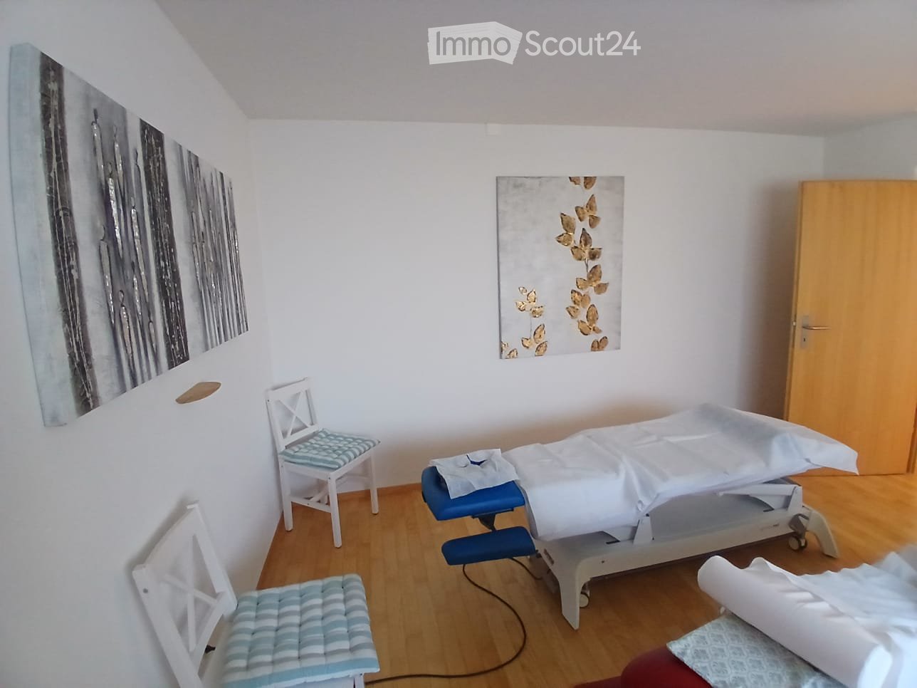 A small, well-lit room with a treatment table, two chairs, and artwork on the walls.