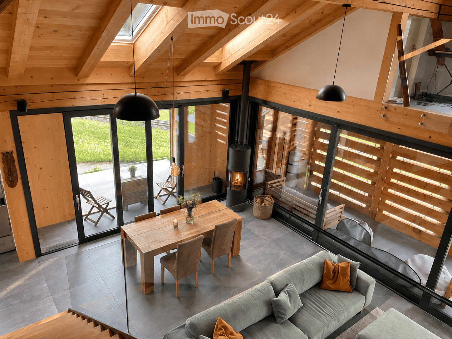 Open-concept living and dining area with wooden beams, large glass doors leading to a patio, a grey sofa with orange pillows, a wooden dining table with chairs, and a modern wood-burning stove.