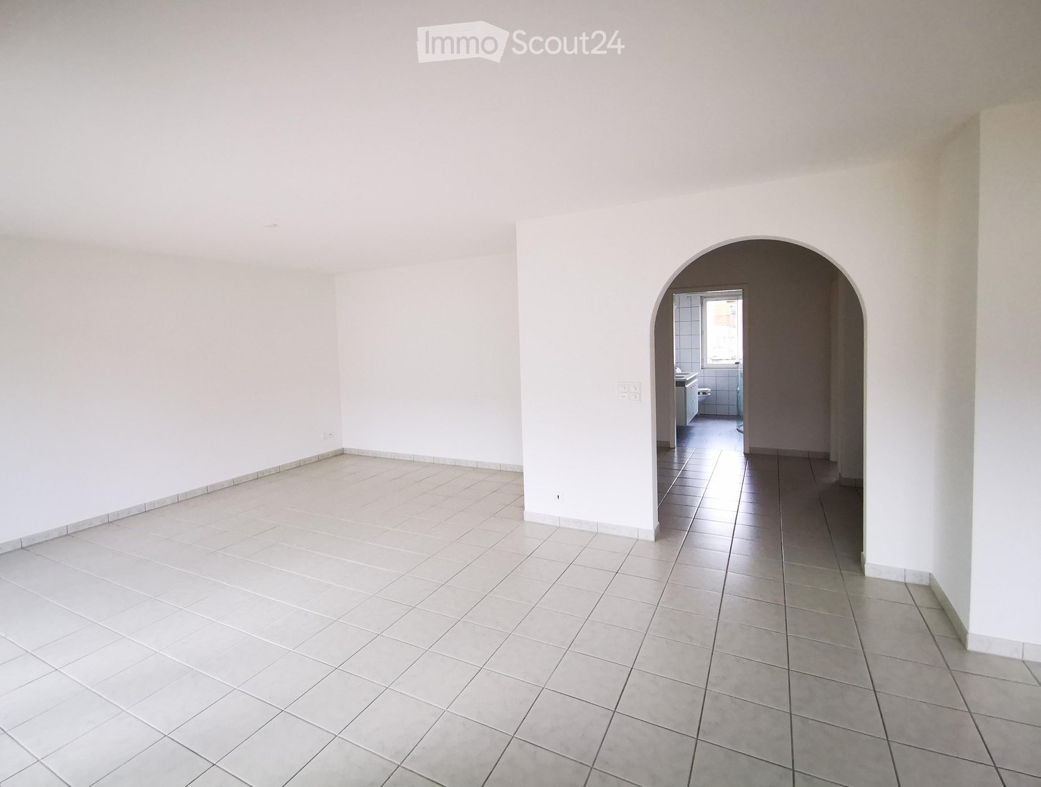 Empty room with white tiled floor and walls, featuring an arched doorway leading to a small bathroom with white tiles and a sink.