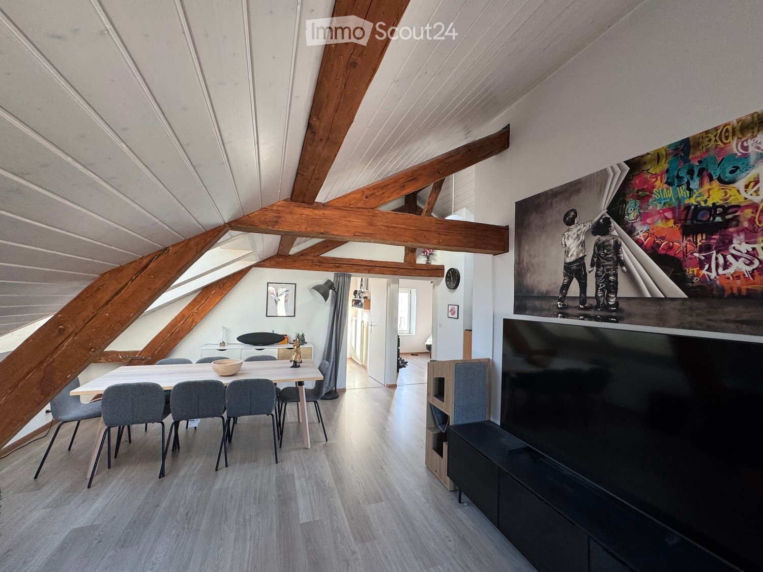 Open-plan living and dining area with exposed wooden beams, modern furniture, and a large screen with a graffiti-style artwork above it.