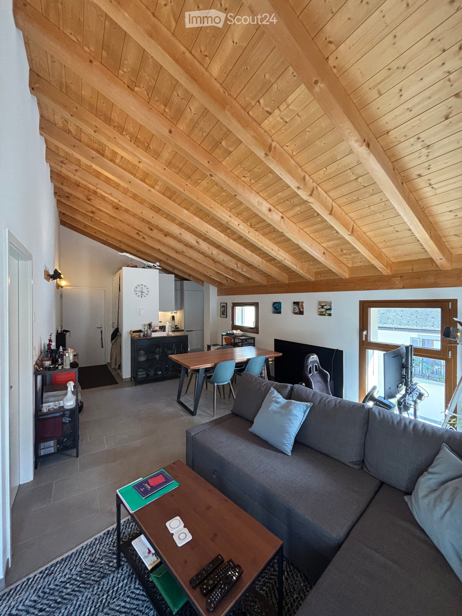 A modern living room with a grey sectional sofa, wooden coffee table, dining table with chairs, and a kitchen area in the background. The room features exposed wooden beams on the ceiling and large windows.