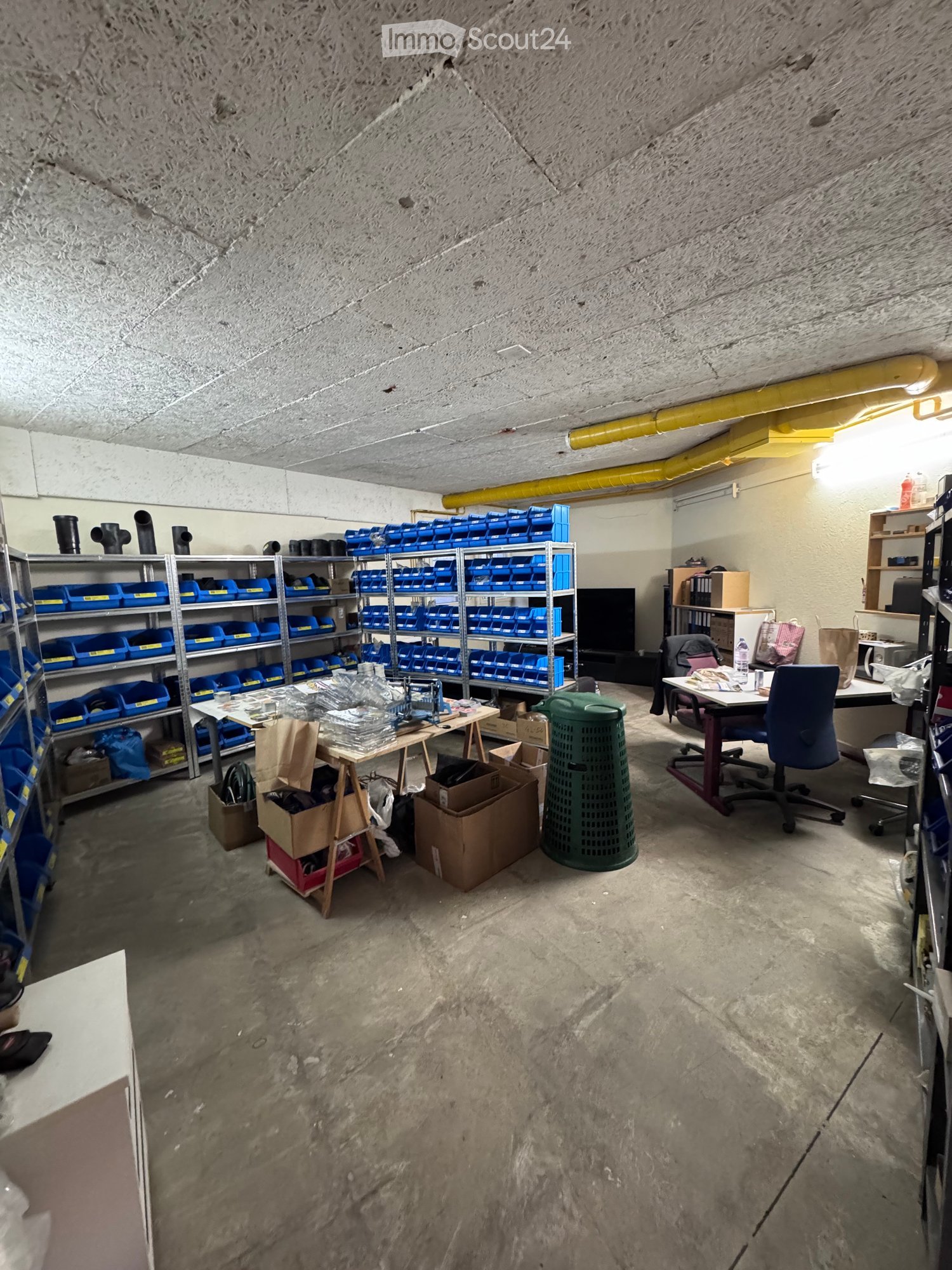 A storage or workshop area with metal shelving units filled with blue storage bins, a desk with a chair, a small table with various items, and a green trash can. The space has a concrete floor and exposed pipes on the ceiling.