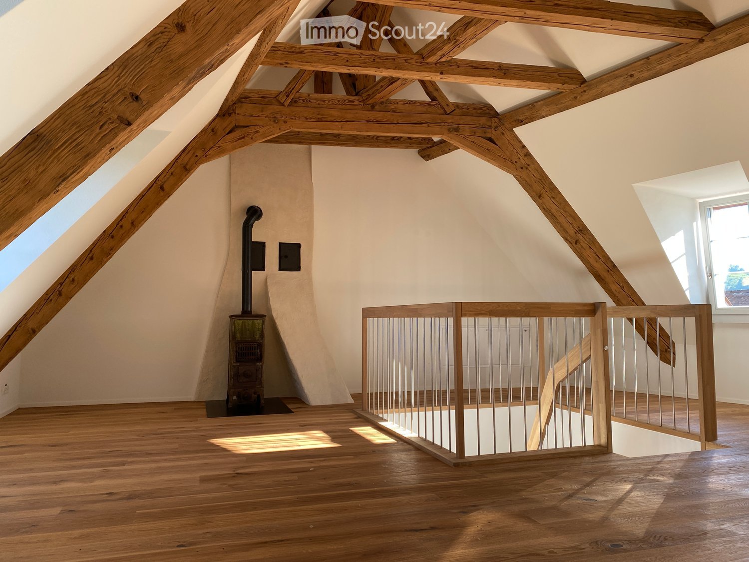 Spacious attic room with exposed wooden beams, wooden flooring, skylights, and a staircase railing. A wood-burning stove is visible against the wall.