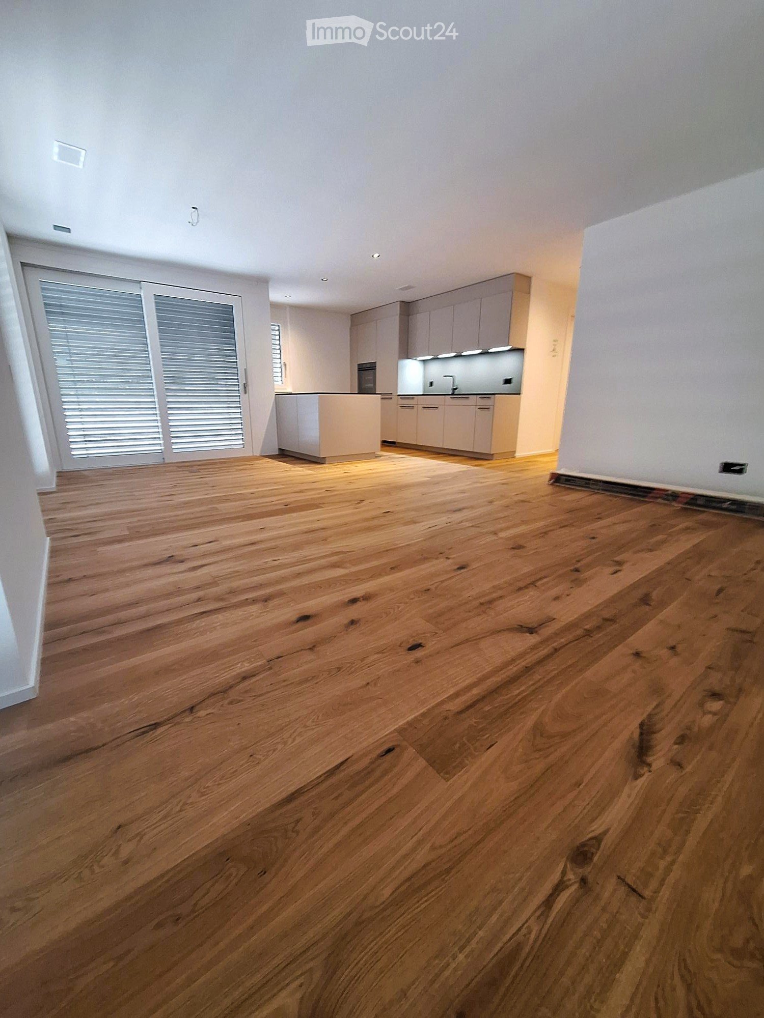 Empty room with polished wooden flooring, white walls, and a small kitchen area with cabinets and a countertop visible in the background.