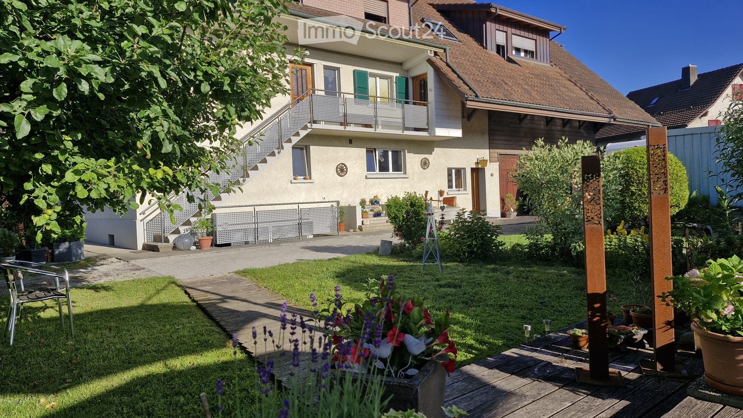 Well-maintained garden with a wooden deck, potted plants, and a metal sculpture. The house features a balcony, multiple windows, and a staircase leading to the entrance.