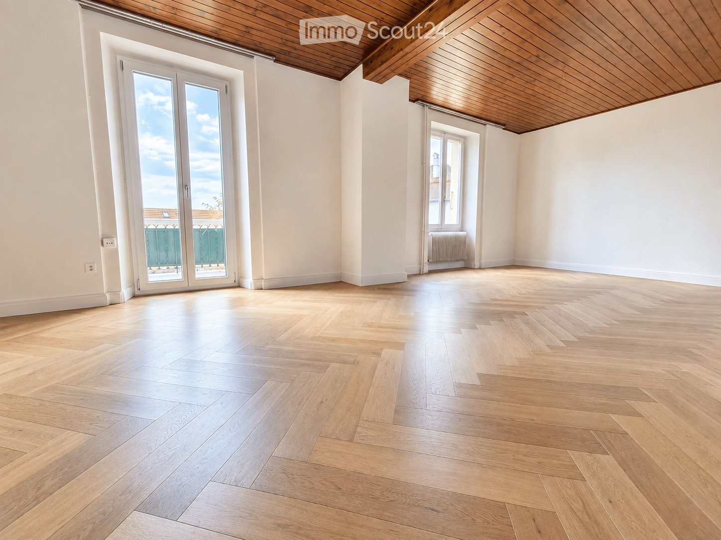 Empty room with light wooden herringbone flooring, white walls, and a wooden ceiling. Features include a large sliding glass door leading to a balcony and a smaller window with a radiator below.