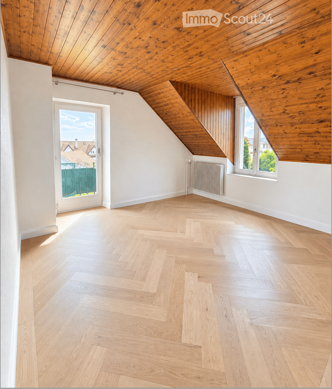 Empty attic room with wooden herringbone flooring, white walls, exposed wooden ceiling beams, and large windows offering natural light and a view of the neighborhood.