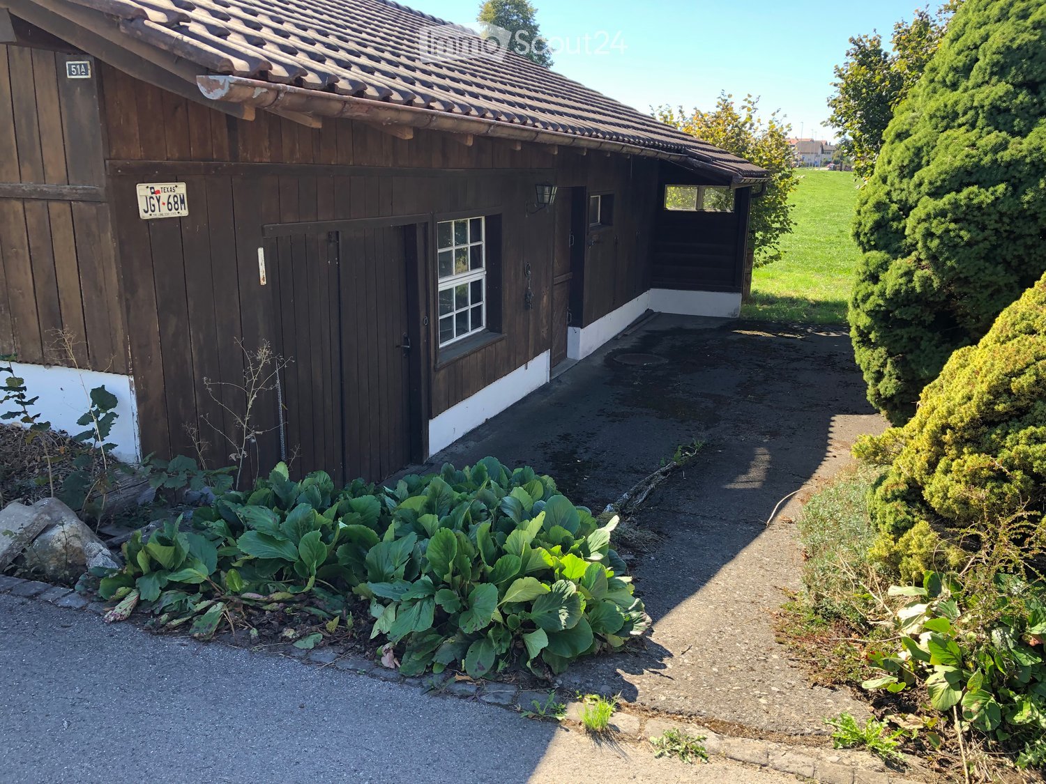 A rustic wooden building with a tiled roof, white baseboards, and a small garden area with leafy plants in front.