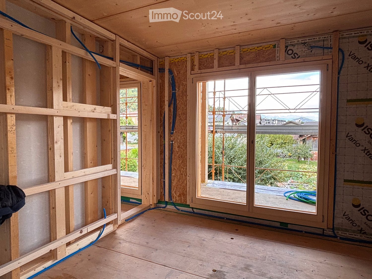 Empty room with exposed wooden framing, unfinished walls, and large windows showing a view of the outdoors.