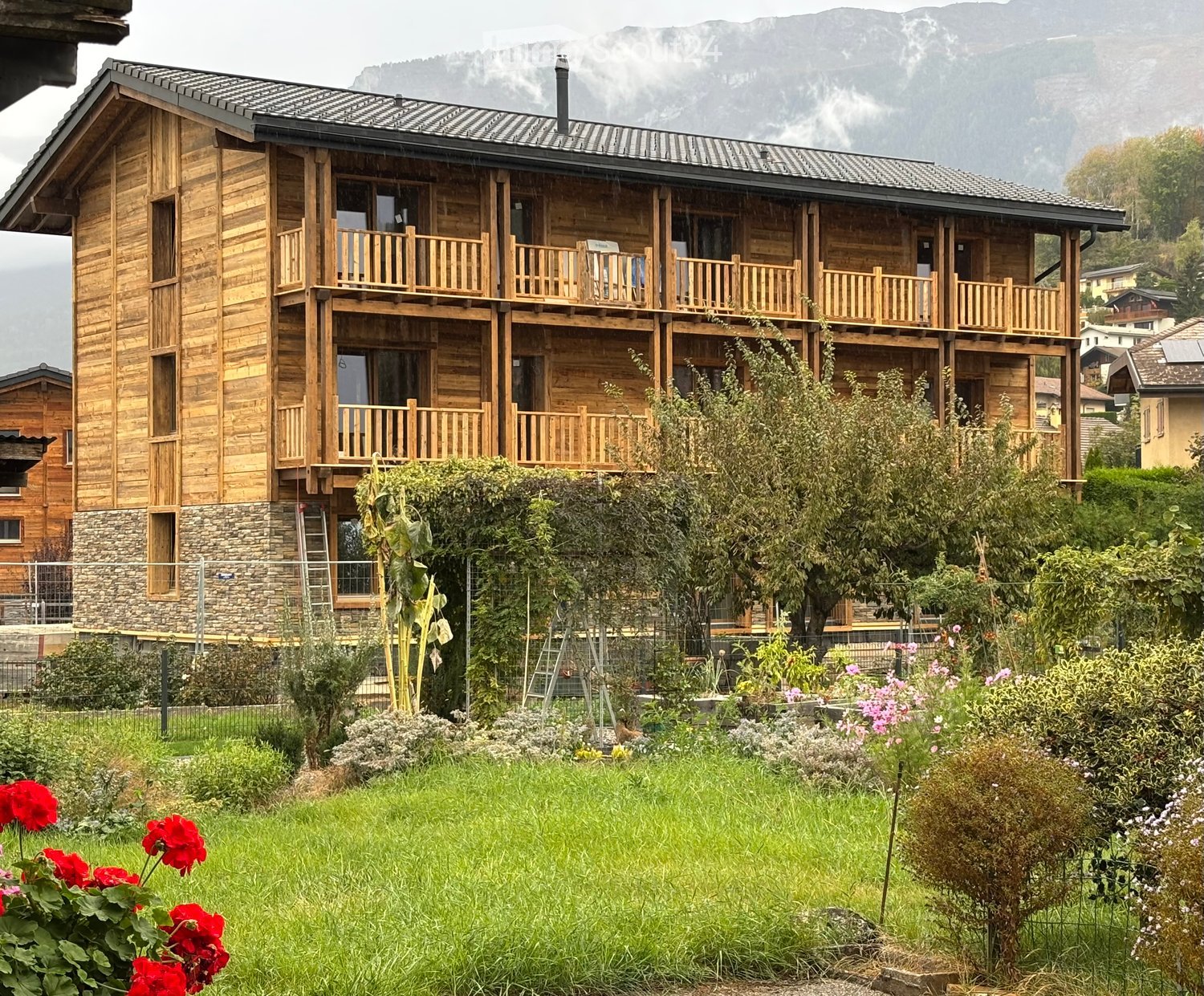 A multi-story wooden building with balconies, surrounded by a garden with flowers and trees, with mountains in the background.